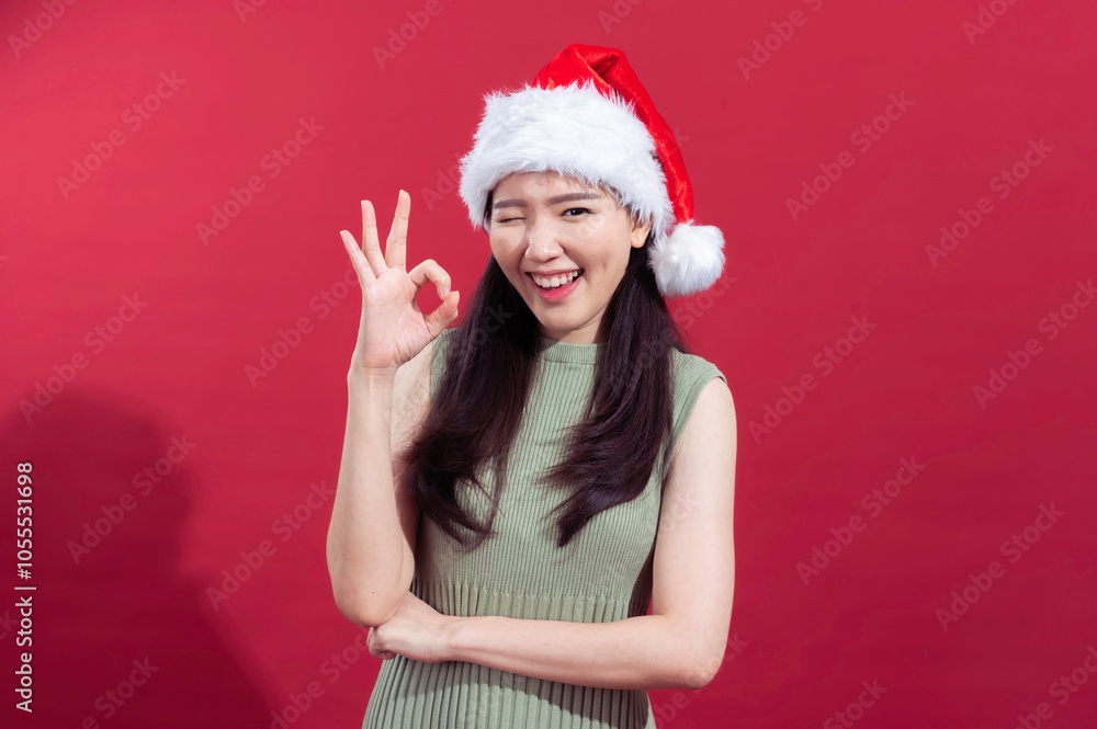 Asian woman in a Santa hat, wearing a green dress, standing against a red background with a cheerful expression, winking and showing an Asian woman in a Santa hat, wearing a green dress, standing against a red background with a cheerful expression, winking and showing an