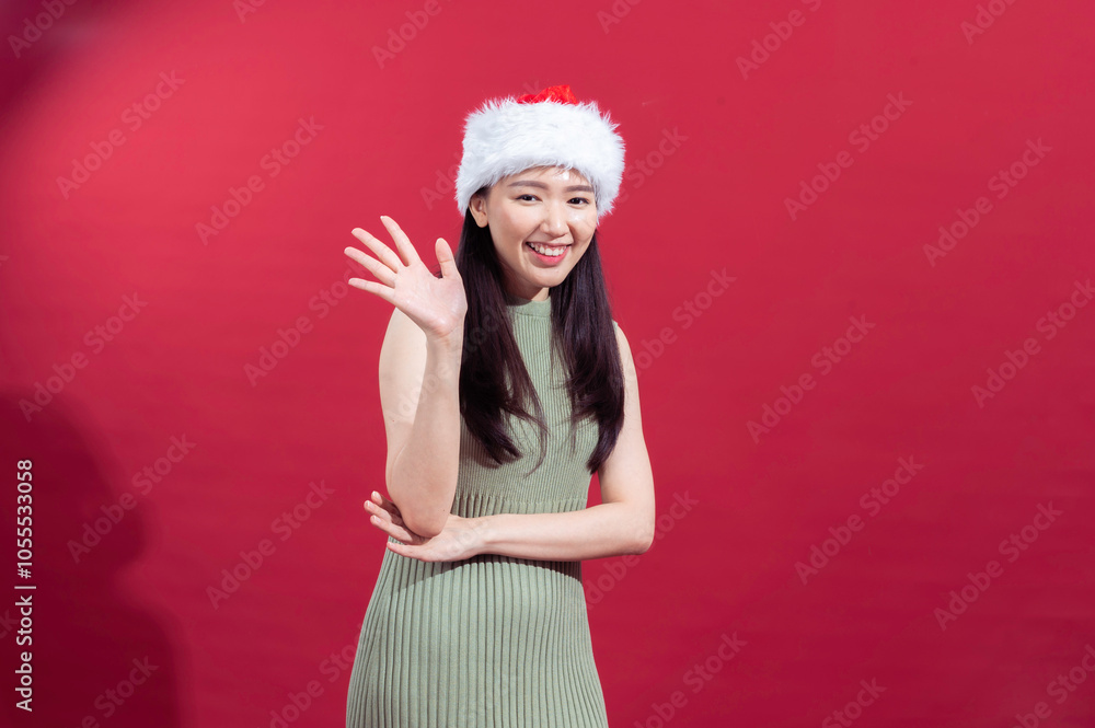 Asian woman in a green dress and Santa hat waving with a cheerful smile, set against a red background, evoking a festive and welcoming holiday spirit Asian woman in a green dress and Santa hat waving with a cheerful smile, set against a red background, evoking a festive and welcoming holiday spirit