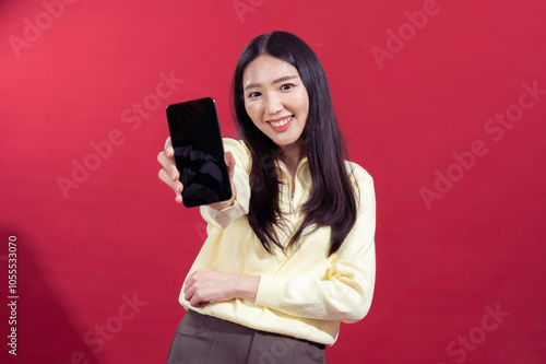 Asian woman smiling while extending a smartphone towards the camera, wearing a yellow sweater against a red background. The focus on the phone suggests a concept related