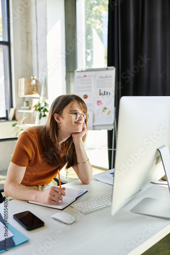 A young non binary person is deep in thought while working at a desk in an inviting office space. photo