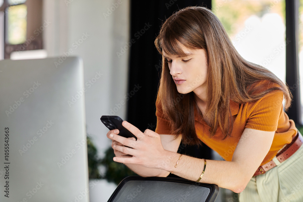 A young non binary individual checks their smartphone while leaning on an office desk. A young non binary individual checks their smartphone while leaning on an office desk.