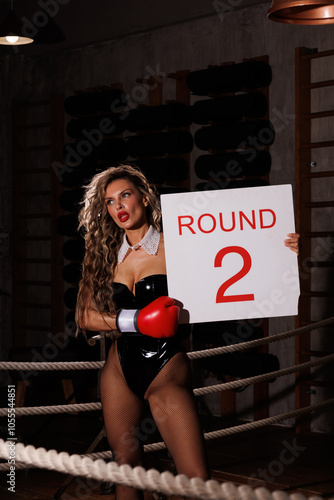 A beautiful girl in red sports gloves. A curly-haired girl in a latex black lacquer bodysuit and fishnet tights in a boxing ring with a punching bag. A woman in the ring shows round 2 with a sign