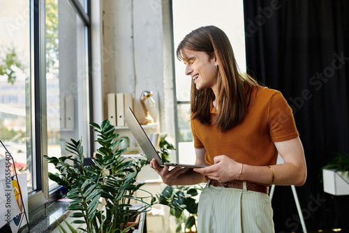 A young non binary person smiles while working on a laptop, surrounded by plants in a bright office. photo