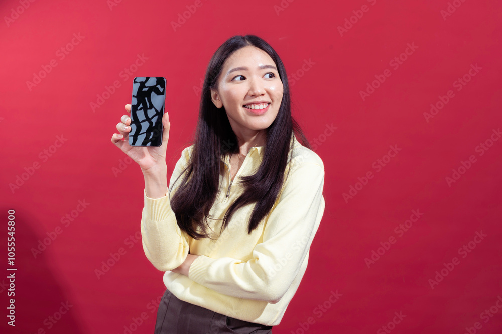 Asian woman smiling while extending a smartphone towards the camera, wearing a yellow sweater against a red background. The focus on the phone suggests a concept related Asian woman smiling while extending a smartphone towards the camera, wearing a yellow sweater against a red background. The focus on the phone suggests a concept related