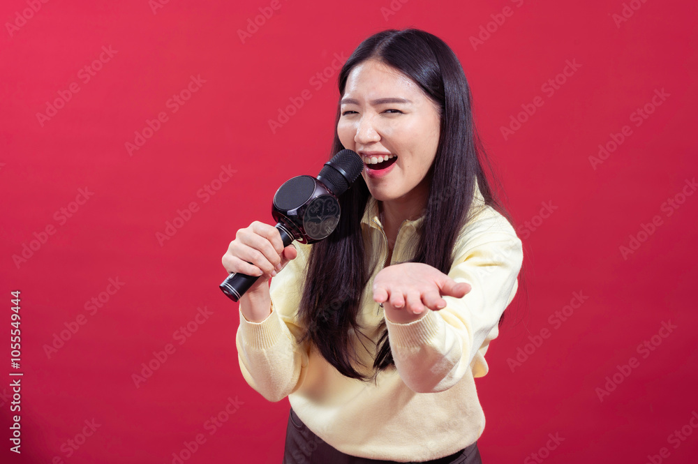 Asian woman smiling and singing into a microphone, wearing a yellow sweater with a red background. She appears enthusiastic and expressive, holding the microphone Asian woman smiling and singing into a microphone, wearing a yellow sweater with a red background. She appears enthusiastic and expressive, holding the microphone