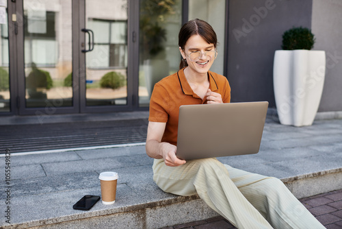 A non binary individual sits on the steps, engaged with a laptop while enjoying coffee. photo