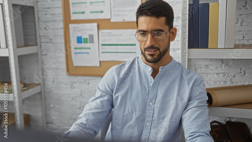 Handsome young man with beard working in modern office interior looking focused at computer screen amidst documents and shelves.