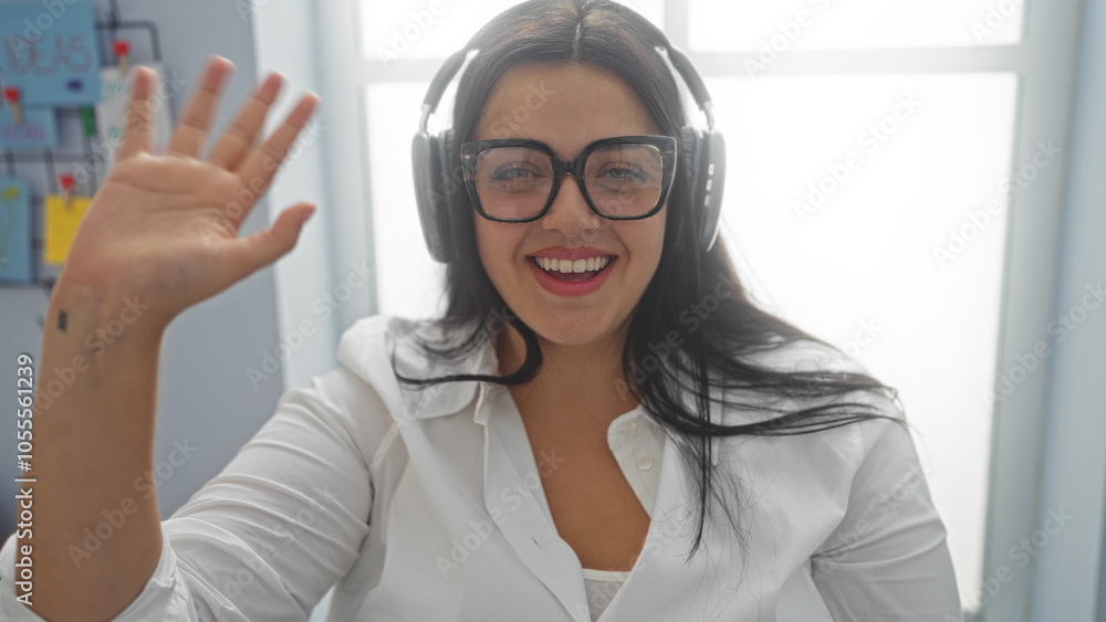 Woman waving with glasses and headphones in an office room smiling and wearing a white shirt Woman waving with glasses and headphones in an office room smiling and wearing a white shirt