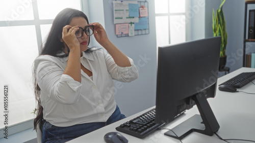 Fotografie Woman adjusting glasses seated at a desk in an office, focusing on a computer screen with motivational notes on the wall behind her, creating a professional indoor workplace setting
