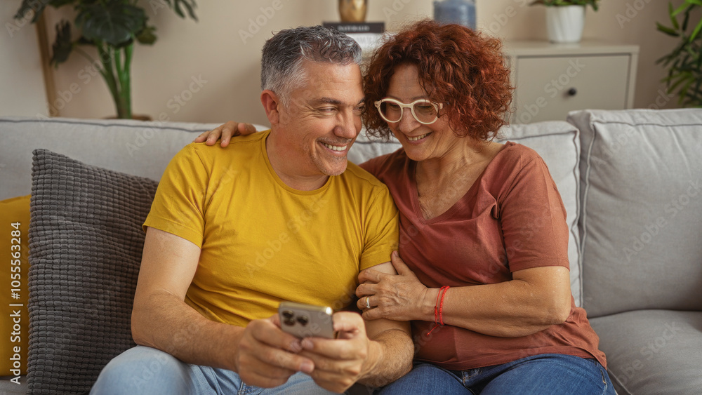 Middle-aged couple sitting together on a couch in a living room smiling and looking at a smartphone in a cozy home interior. Middle-aged couple sitting together on a couch in a living room smiling and looking at a smartphone in a cozy home interior.