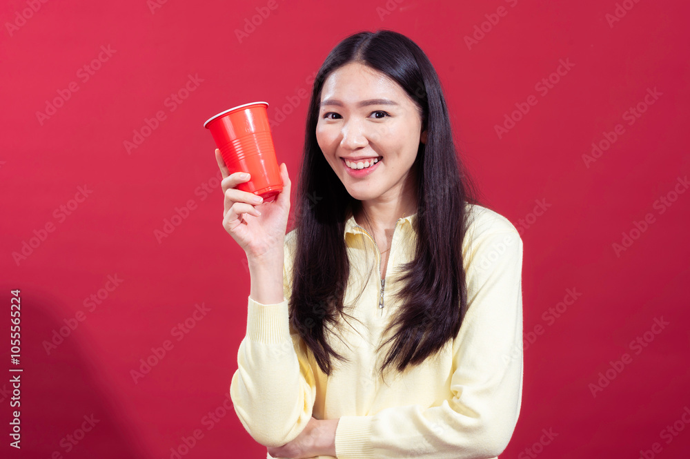 Asian woman smiling and holding a red plastic cup in her hand, wearing a yellow sweater against a red background. She looks cheerful and relaxed, with one arm crossed and the other holding cup Asian woman smiling and holding a red plastic cup in her hand, wearing a yellow sweater against a red background. She looks cheerful and relaxed, with one arm crossed and the other holding cup