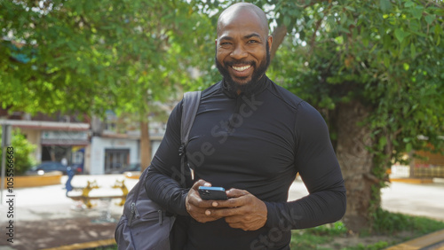 Handsome man standing outdoors in a city park, smiling while holding a smartphone and carrying a backpack, with trees and urban buildings in the background