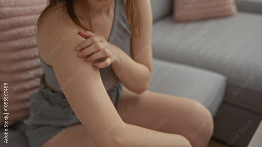 Young woman scratching her shoulder while sitting on a couch in a living room at home wearing casual clothes Young woman scratching her shoulder while sitting on a couch in a living room at home wearing casual clothes