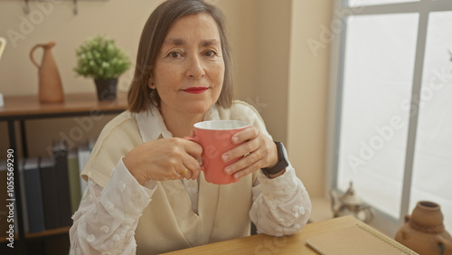 Mature hispanic woman with short hair enjoying coffee indoors at home, near a window, conveying tranquility.