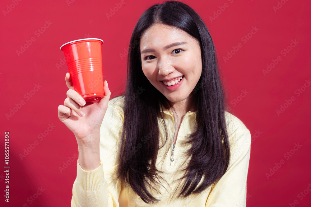 Asian woman smiling and holding a red plastic cup in her hand, wearing a yellow sweater against a red background. She looks cheerful and relaxed, with one arm crossed and the other holding cup Asian woman smiling and holding a red plastic cup in her hand, wearing a yellow sweater against a red background. She looks cheerful and relaxed, with one arm crossed and the other holding cup