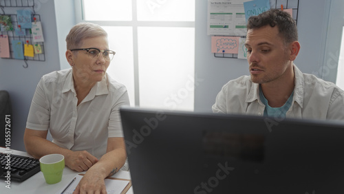 Woman and man, both adults, working together on a project in a modern office, with documents and a computer screen in a professional workplace setting.