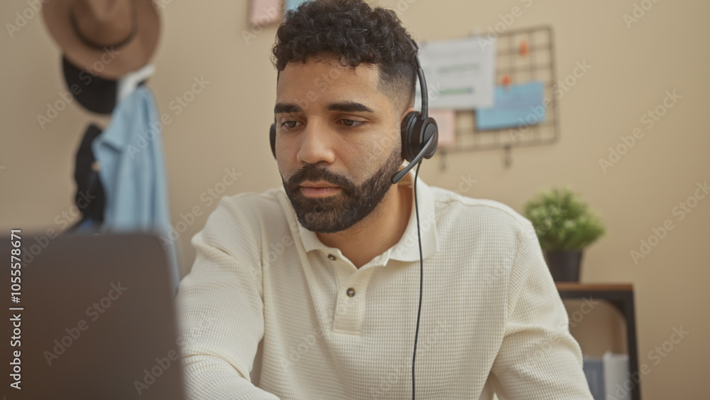 A focused hispanic man with a beard works remotely from his apartment using a laptop while wearing a headset. A focused hispanic man with a beard works remotely from his apartment using a laptop while wearing a headset.