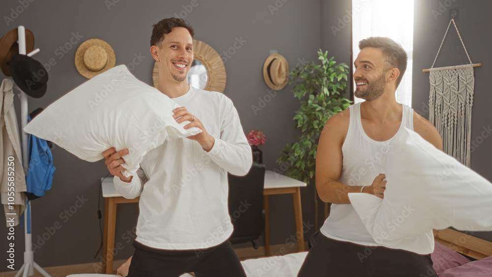 Fototapeta premium Men smiling while having a playful pillow fight on the bed in a cozy bedroom adorned with hats and wall decor, capturing a moment of love and joy in a home setting.