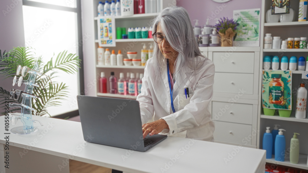 Middle aged woman with grey hair and glasses working on a laptop in a pharmacy, surrounded by medicine bottles, eyeglasses, and plant in the interior store setting. Middle aged woman with grey hair and glasses working on a laptop in a pharmacy, surrounded by medicine bottles, eyeglasses, and plant in the interior store setting.