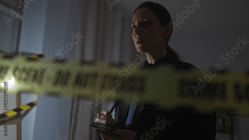 A serious female officer stands at an indoor crime scene with 'do not cross' tape and notepad. photo