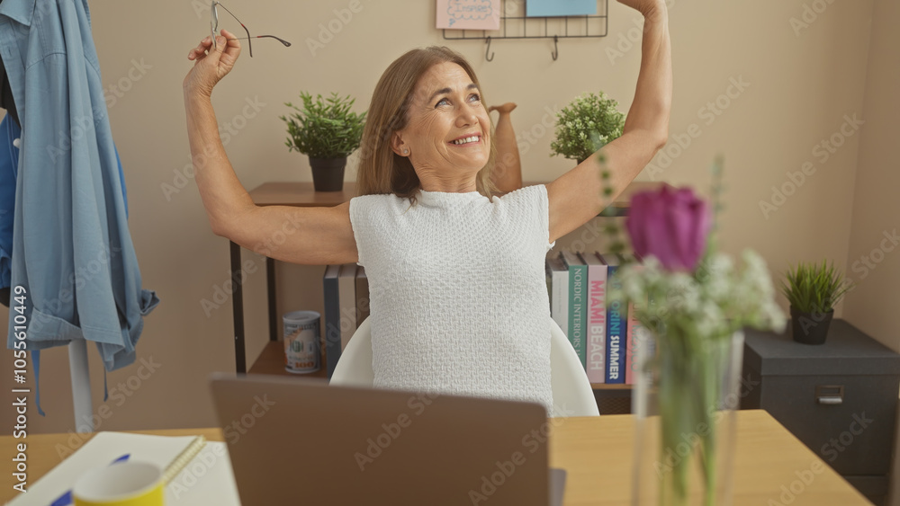 Mature woman stretching arms with a smile, relaxed ambiance in a home office with books, flowers, and laptop. Mature woman stretching arms with a smile, relaxed ambiance in a home office with books, flowers, and laptop.