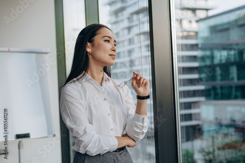 Young businesswoman stands by office window gazing thoughtfully at city skyline