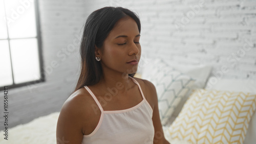 Young woman sitting on a bed in a bedroom with eyes closed and breathing peacefully, showcasing serene indoor moments in her home. Free Download Young woman sitting on a bed in a bedroom with eyes closed and breathing peacefully, showcasing serene indoor moments in her home. - GFXLoad
