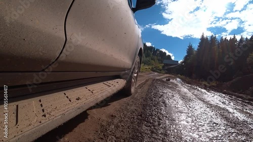 Black car driving on muddy road in mountains. Road is bumpy, and there are stones on side. Driver carefully navigates in difficult terrain. View of camera from roof of car.