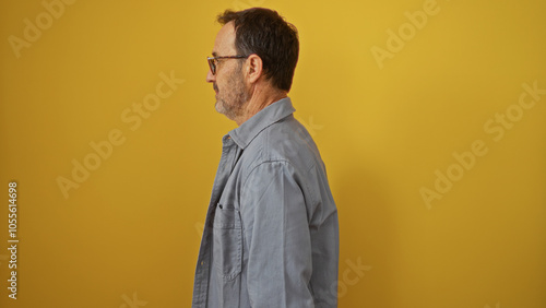 Middle-aged hispanic man in blue shirt standing sideways against isolated yellow background