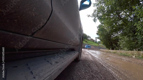 Black car driving on muddy road in mountains. Road is bumpy, and there are stones on side. Driver carefully navigates in difficult terrain. View of camera from roof of car.