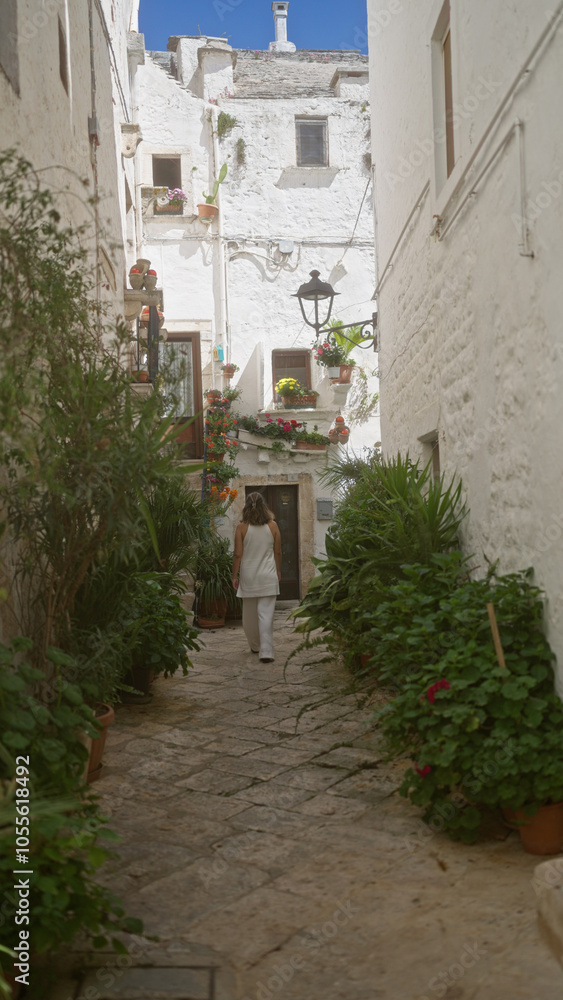Fototapeta premium Young woman walking through the charming white streets of locorotondo, puglia, italy, adorned with vibrant plants and flowers in the old town on a sunny day.