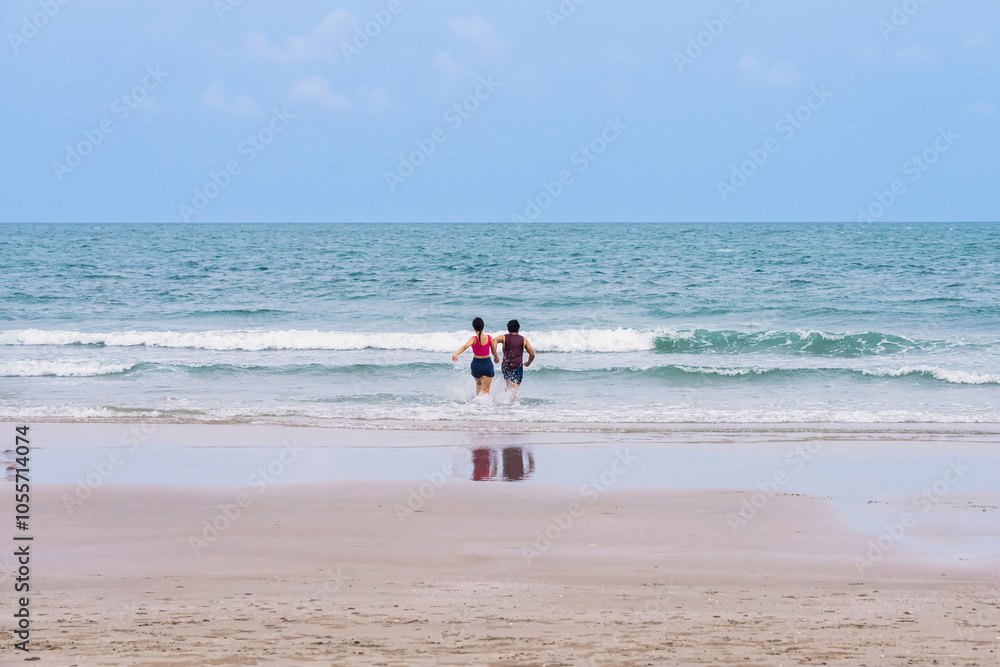 Back view of happy carefree couple holding hands and running in water at beach. Couple plays in ocean waves. Happy young couple playing in sea water. Guy and girl splashing together, beach vacation. Back view of happy carefree couple holding hands and running in water at beach. Couple plays in ocean waves. Happy young couple playing in sea water. Guy and girl splashing together, beach vacation.