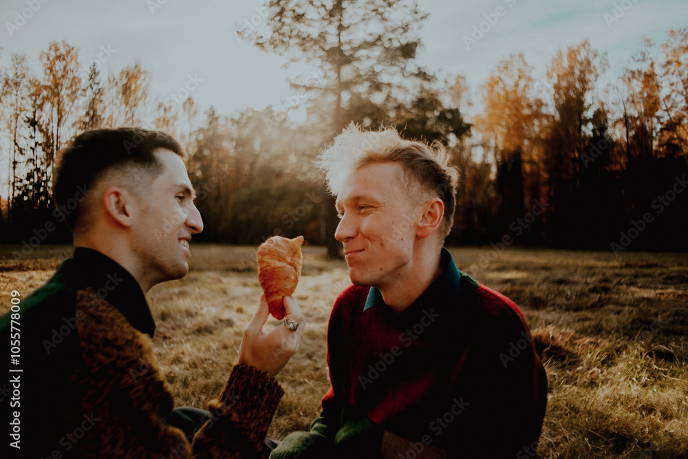 Obraz premium On a sunny autumn day, two friends have a picnic in the countryside. One friend feeds the other a croissant, and they enjoy each other's company. They feel happy and loved.