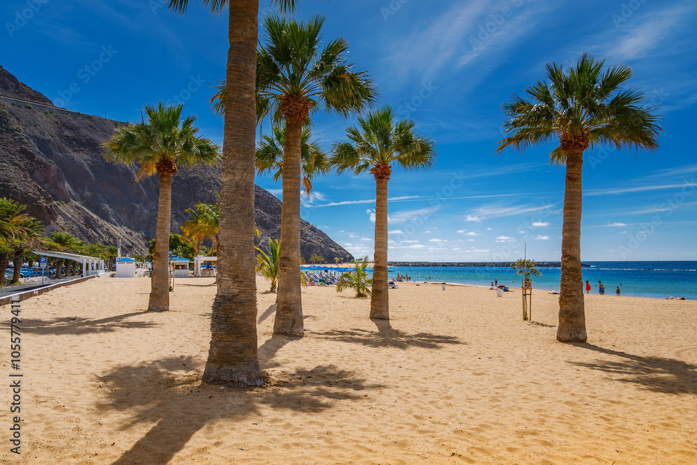 A view of Las Teresitas beach in Tenerife, showcasing golden sand, palm trees, and a calm blue ocean with mountains in the background A view of Las Teresitas beach in Tenerife, showcasing golden sand, palm trees, and a calm blue ocean with mountains in the background