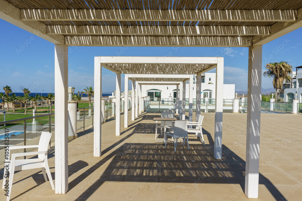 Fototapeta premium Relaxing terrace view overlooking the ocean with stylish seating and palm trees under a sunny sky.