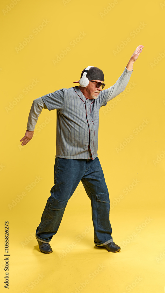 Full-length image of elderly bearded man in cap and sunglasses listening to music in headphones and dancing against yellow background. Concept of human emotions, elderly lifestyle. Copy space, ad Full-length image of elderly bearded man in cap and sunglasses listening to music in headphones and dancing against yellow background. Concept of human emotions, elderly lifestyle. Copy space, ad