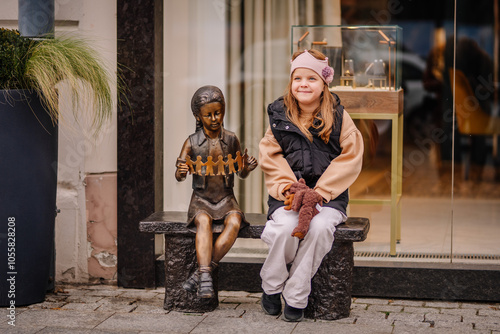 A young girl sits next to a bronze statue of a child holding paper cutouts of a family. The scene is outside a shop window with greenery around.. photo