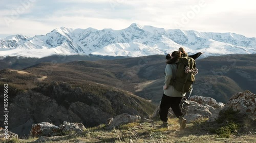 Back view at two hiker women with backpack raising arms for joy, celebrating climbing success standing on high mountain peak at scenic landscape. Travel achievements and recreation together