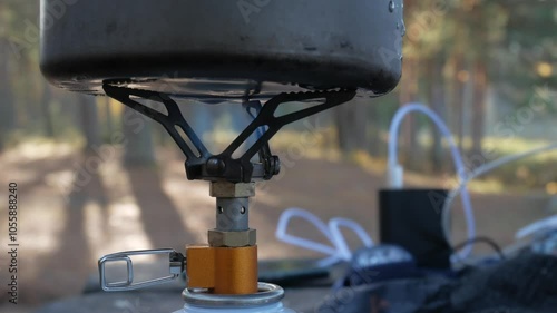 Camping pot on a lit gas burner, cooking food outdoors in a forest setting.