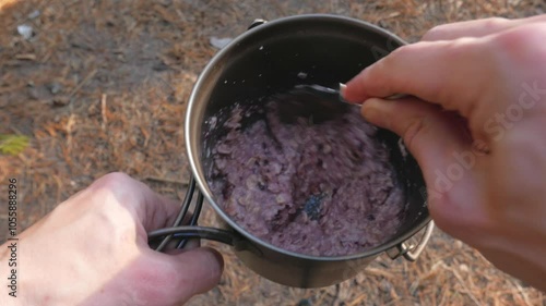 Outdoor camp breakfast of berry oatmeal, stirred with a spoon in a pot, first-person view.