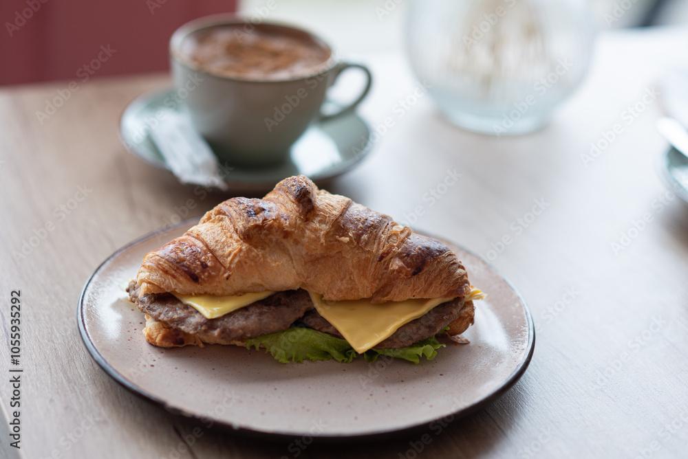 Close-up of a mug with coffee and a croissant with cheese and ham standing on a table in a cafe, near the window. Close-up of a mug with coffee and a croissant with cheese and ham standing on a table in a cafe, near the window.
