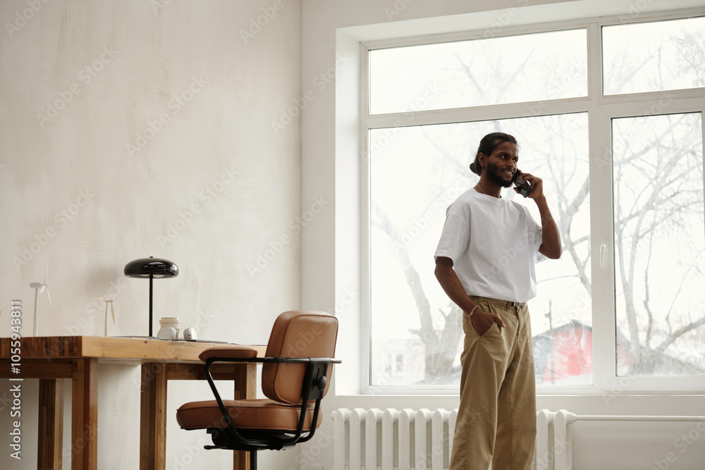 Architect standing near large window while talking on phone in bright room with wooden desk and chair Architect standing near large window while talking on phone in bright room with wooden desk and chair