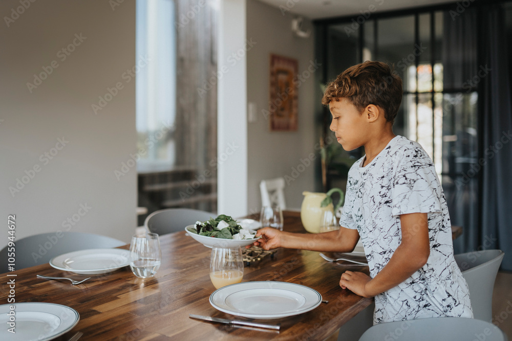 Cute boy keeping salad bowl while setting dining table for lunch at home Cute boy keeping salad bowl while setting dining table for lunch at home