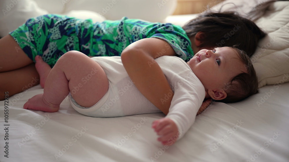 Older sibling cuddling baby on bed, both lying together, with baby smiling and looking upward while the sibling rests beside, showing warmth and love Older sibling cuddling baby on bed, both lying together, with baby smiling and looking upward while the sibling rests beside, showing warmth and love