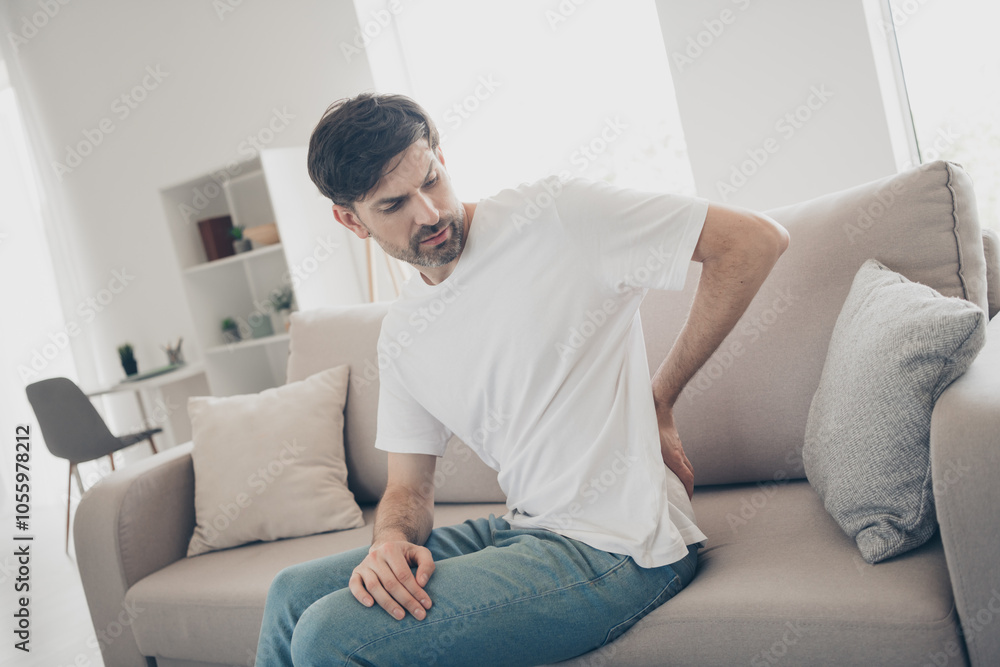 Young man experiencing back pain while sitting on a couch in a bright living room interior, highlighting discomfort and health awareness Young man experiencing back pain while sitting on a couch in a bright living room interior, highlighting discomfort and health awareness