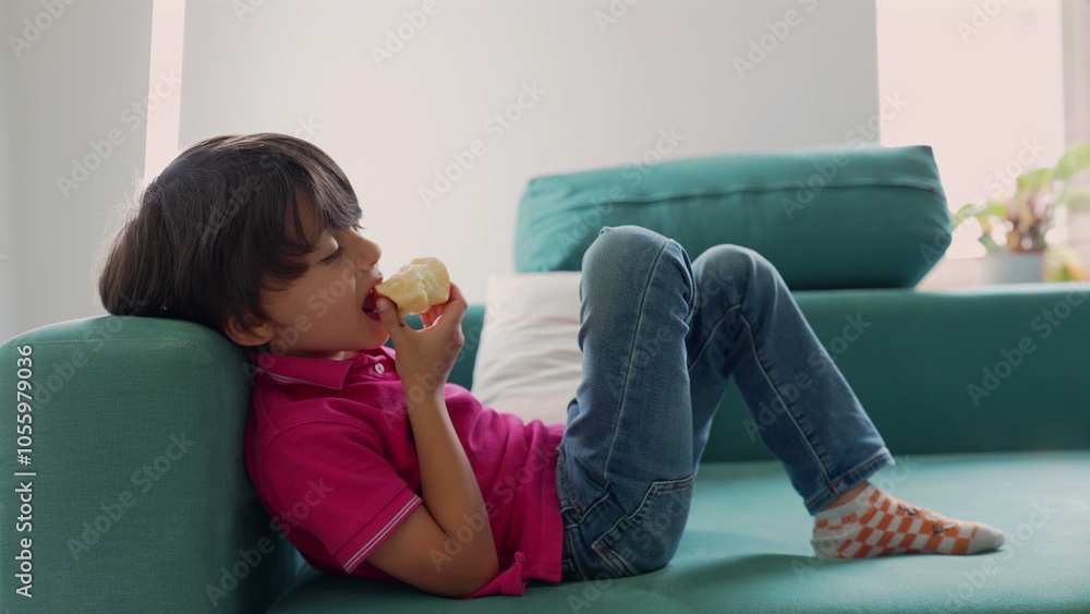 Young boy relaxing on couch while eating an apple, leaning against the sofa and appearing thoughtful, in a calm and quiet indoor setting Young boy relaxing on couch while eating an apple, leaning against the sofa and appearing thoughtful, in a calm and quiet indoor setting