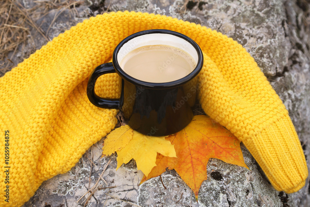 Hot milk coffee in enamel black mug standing on gray stone, yellow scarf and autumn leaves as decor. Fall hygge mood. Outdoors, picnic, camping, travel, trekking concept.