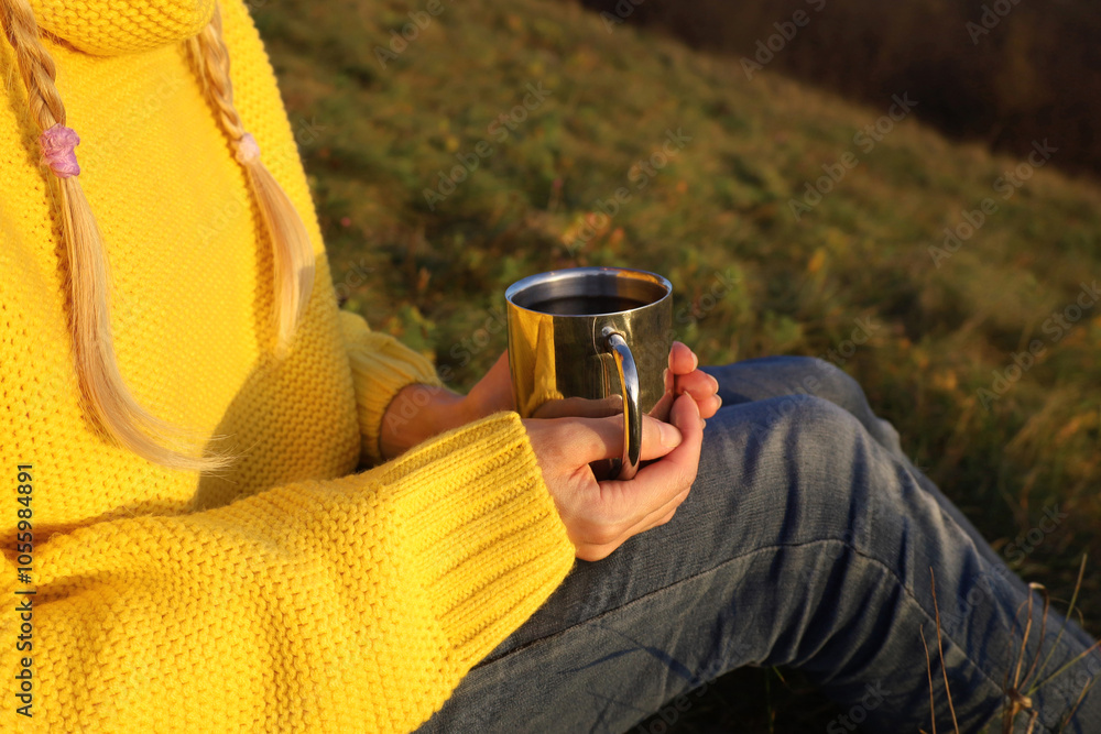Blonde woman in yellow sweater and blue jeans holds in hands stainless steel cup with hot tea. Autumn hygge mood. Outdoors, picnic, camping, travel, trekking concept. No face, unrecognizable.