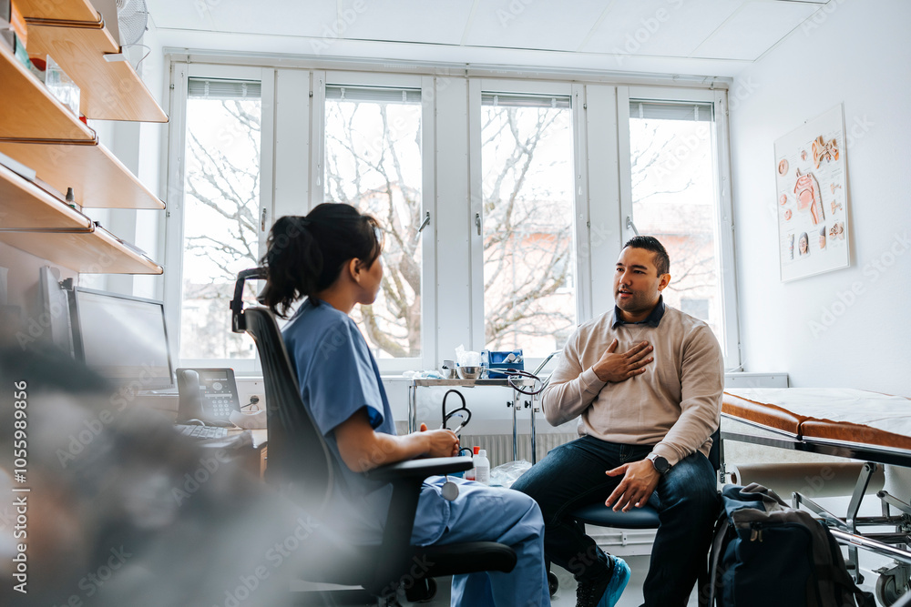 Man with hand on chest talking with female medical expert while sitting in examination room Man with hand on chest talking with female medical expert while sitting in examination room