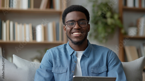 A realistic portrait of a young man exuding confidence in a comfortable, well-decorated living room. Heâs seated with a relaxed posture, holding a tablet and dressed in a casual bl photo